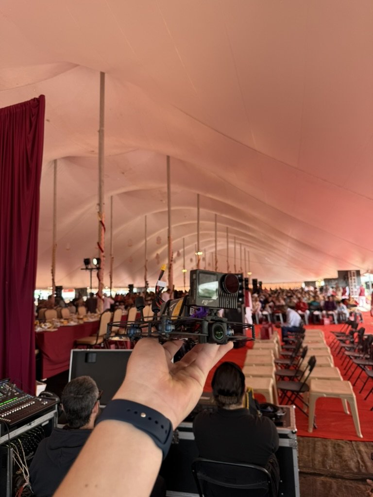 A ducted FPV cinewhoop held up inside an event marquee before a shoot, with rows of seating and the stage behind it
